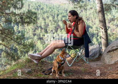 Schwangere Frau sitzt in einem Campingstuhl, isst einen Snack aus einer Tasse mit ihrem Hund an ihrer Seite und genießt den Waldblick Stockfoto