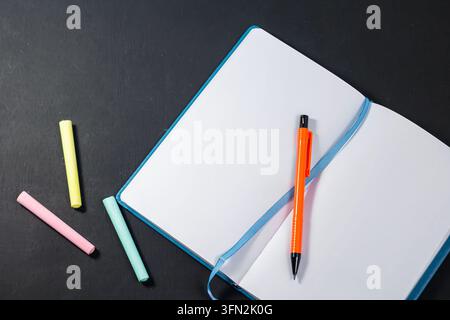Ein offenes leeres blaues Notizbuch mit einem hellen orangefarbenen Bleistift darin, das neben bunten Kreidestücken auf einer dunklen Holzoberfläche platziert ist, perfekt für zurück zur Schule Konzepte. Hochwertiges Foto Stockfoto