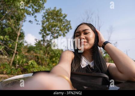 Junge stilvolle asiatische Frau, die sich aus dem Sonnendach eines Autos lehnt und ein Selfie-Porträt macht. Stockfoto