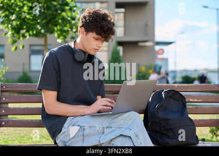 Konzentrierter junger männlicher Student, der im Freien auf einer Holzbank einen Laptop benutzt Stockfoto