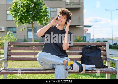 Junger männlicher Student hört Musik mit Kopfhörern und sitzt auf einer Bank im Freien im Park Stockfoto