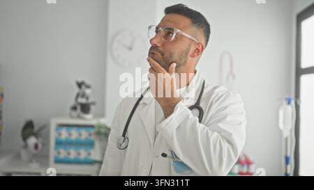 Männerarzt trägt weißen Kittel und klare Brille mit Stethoskop und Hand am Kinn im Untersuchungsraum der Klinik; Nachdenken. Stockfoto
