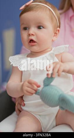 Kaukasisches Baby-Mädchen greift nach einem gestrickten Hasen, die Hand umklammert den Plüsch, während die Mutter ihre Hüften und ihren Schoß im Studio-Setting mit blauer Wand unterstützt; Kuriosität Stockfoto