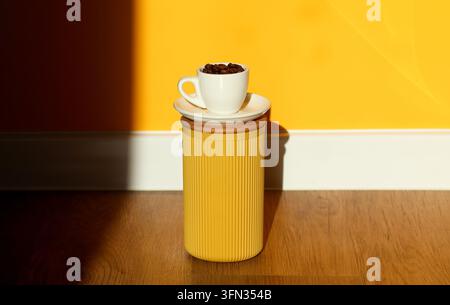 Eine weiße Tasse gefüllt mit gerösteten Kaffeebohnen auf einer Untertasse auf einem gelben zylindrischen Behälter vor einem lebendigen gelb-schwarzen Wandhintergrund. Minima Stockfoto