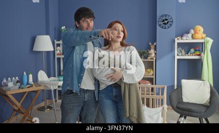 Frau, die ein in eine Decke gewickeltes Neugeborenes in einem blauen Kinderzimmer hält, mit einem lächelnden Mann neben einem Holzbett und einem Wickeltisch; Zärtlichkeit. Stockfoto