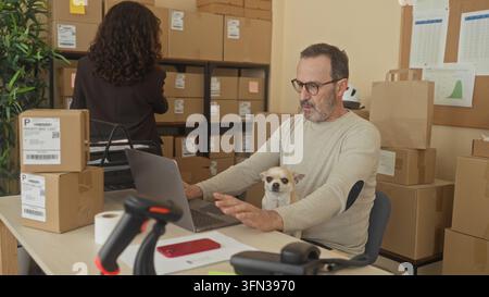 Mann tippt auf Laptop mit kleinem Hund auf dem Schoß und gestapelten Versandpaketen, scannt Etiketten mit Barcodeleser im Gebäude; konzentrierte Produktivität. Stockfoto