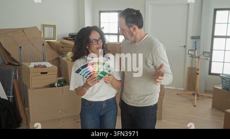 Mittelaltes Paar hält einen Pinselventilator, während die Frau auf einen Farbmuster zeigt, der Mann gestikuliert, umgeben von Kartons und Arbeitsleuchten in einem Gebäude; Haus im Stockfoto