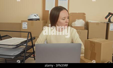 Frau am Schreibtisch sortiert Pakete und Papierkram neben einem Laptop, greift nach einem Dokumentenbehälter und hält die Hand an den Hals in einem Verpackungsgebäude; E-Commerce-Erschöpfung Stockfoto