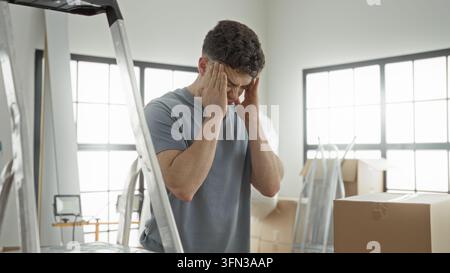 Mann drückt die Hände an die Schläfen neben gestapelten Kartons und einer Metallleiter im Gebäude beim Auspacken von Paketen; Stress überwältigt Bewegungsmüdigkeit. Stockfoto