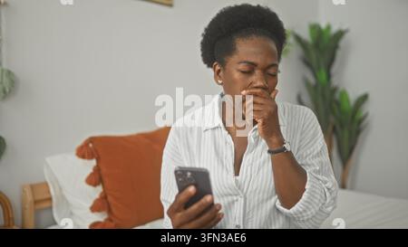 Mittlere Altersfrau auf dem Bett, die ein Smartphone hält, mit der Hand am Hals und hustend, Augen geschlossen, weißes Hemd und orangefarbenes Kissen sichtbar im Gebäude; Gesundheit c Stockfoto