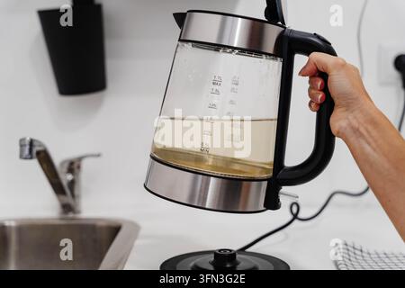 Eine Hand hebt einen gläsernen Wasserkocher, der mit trübstem gelbem Leitungswasser gefüllt ist, über ein Küchenwaschbecken. Entfärbtes Wasser wie dieses veranschaulicht Trinkwasser Stockfoto
