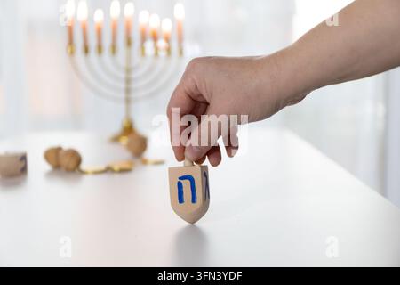 Nahaufnahme einer Hand, die einen hölzernen Dreidel hält, bereit zum Drehen, auf einem weißen Chanukka-Tisch mit einer sanft beleuchteten Menora im Hintergrund, die Wärme ausstrahlt Stockfoto