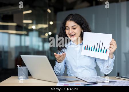 Lächelnde Geschäftsfrau führt eine virtuelle Konferenz auf einem Laptop, zeigt ein Balkendiagramm auf Papier, diskutiert über Geschäftsanalysen und erreicht Erfolg während der Telearbeit im Büro Stockfoto
