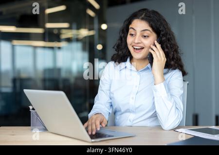 Junge Frau in einem modernen Büro, die ein fröhliches Gespräch an ihrem Mobiltelefon führt, während sie auf einem Laptop tippt, und Erfolg, Kommunikation und berufliche Leistung repräsentiert Stockfoto