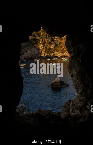 Gerahmte Ansicht aus dem Inneren einer dunklen Küsten-Seenhöhle mit Blick auf Boote auf dem Wasser, die über eine ruhige Mittelmeerlandschaft bei Sonnenuntergang hinausschauen, Isola Bella in Taormina Stockfoto