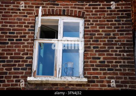 Ein verwittertes weiß gestrichenes Holzfenster in einer roten Backsteinmauer mit einem Keramikkanne auf der Fensterbank im historischen Ribe, Dänemark. Stockfoto