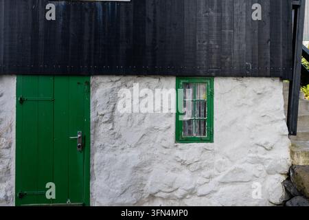 Ansicht eines traditionellen Hauses mit weißen Steinwänden, schwarzer Holzverkleidung, einer grünen Tür und einem grün gerahmten Fenster auf den Färöern. Stockfoto