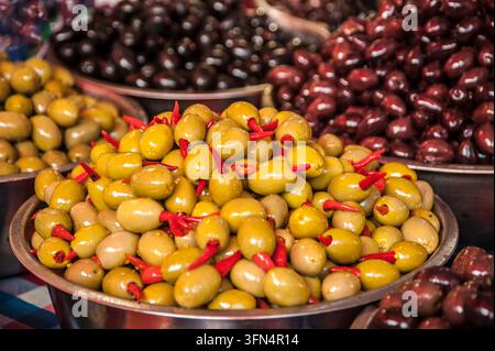 Eine Nahaufnahme von verschiedenen marinierten grünen Oliven, gefüllt mit roten Paprikaschoten und dunklen Oliven, die in Metallschalen auf einem Lebensmittelmarktstand präsentiert werden. Stockfoto