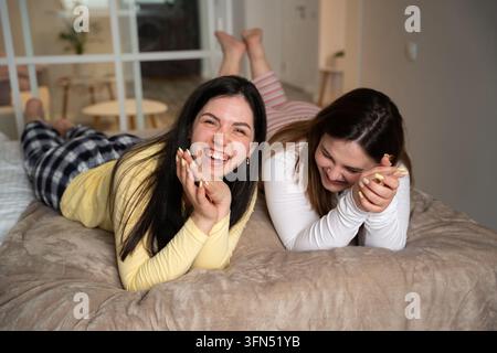 Zwei erwachsene Frauen lachen zusammen, während sie in bequemen Pyjamas zu Hause auf einem Bett liegen. Weibliche Freundschaft, Übernachtung, Spaß und spielerische Freizeitaktivitäten. Stockfoto