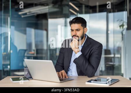 Junger Geschäftsmann im Anzug, der auf einem Holzschreibtisch auf seinen Laptop konzentriert ist, Lösungen überdenkt und Daten in einem modernen Firmenbüro verarbeitet, ernst und fokussiert auf Strategie und Wachstum Stockfoto