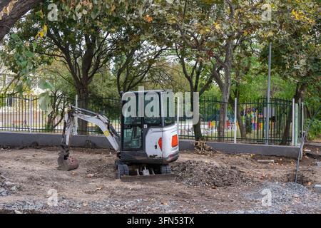 ein weißer Minibagger, der auf einer Schotter- und Kiesbaustelle geparkt ist, umgeben von Bäumen und einem grünen Metallzaun. Stockfoto