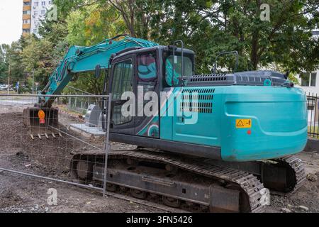 ein blaugrüner Kettenbagger, der hinter einem provisorischen Metall-Sicherheitszaun auf einer Schotter- und Schmutz-Baustelle mit Bäumen und einem Gebäude im Hintergrund geparkt ist Stockfoto