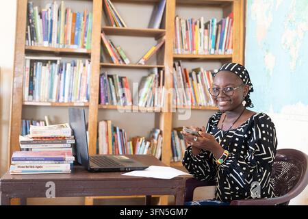 Lächelnde afrikanische Frau verwendet ein Smartphone neben einem Laptop in einer Bibliothek, veranschaulicht digitale Kompetenz, Bildung, berufliche Arbeit, Kommunikation und f Stockfoto