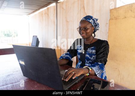 Junge afrikanische Frau arbeitet an einem Laptop an einem Schreibtisch im Freien, trägt ein gemustertes Kopftuch und ein Kleid, illustriert digitale Arbeit, Bildung, Unternehmertum Stockfoto