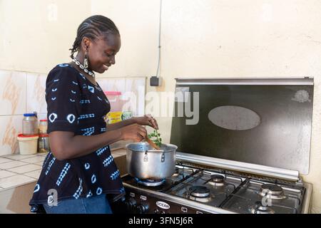 Lächelnde afrikanische Frau kocht eine Mahlzeit auf einem Gasherd in einer Hausküche, rührt in einem Kochtopf mit frischen Kräutern und repräsentiert die Nahrungszubereitung, Ernährung Stockfoto