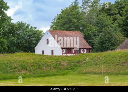Kleines weißes Landhaus mit rotem Dach auf einem grasbewachsenen Hügel, umgeben von grünen Bäumen unter einem bewölkten Himmel. Stockfoto