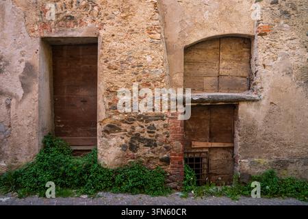 Horizontale Aufnahme einer alten Stein- und Ziegelmauer in Garessio. Die Fassade zeigt zwei historische, zeitlich abgenutzte Holztüren, eine durch einen Balken verbarrikadiert, antik Stockfoto