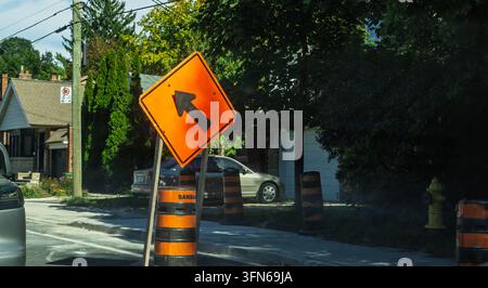 24.09.2026, Toronto, Kanada – Helles orangefarbenes Linksabbiegewarnungsschild und gestreifter Verkehrsfass, die eine Straßenbaustelle in einer Vorstadtstraße markieren Stockfoto