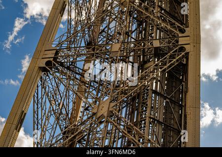 Metallgitter erhebt sich vor blauem Himmel mit Wolken. Stahlträger bilden eine kunstvolle, aufragende Struktur. Sonnenlicht betont Textur und Tiefe des Gerüsts. Stockfoto