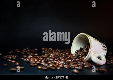 Geröstete Kaffeebohnen, die aus einer Keramiktasse auf eine dunkle Oberfläche mit schwarzem Hintergrund und großzügigem Kopierraum rieseln. Stockfoto