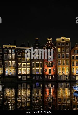 Traditionelle schmale Kanalhäuser bei Nacht beleuchtet mit Spiegelungen auf dem Wasser in Amsterdam, Niederlande. Stockfoto
