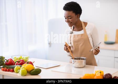Happy Black Hausfrau Kochen Tragen Schürze Browsing Rezepte Online Via Digital Tablet Computer Stehend In Der Modernen Küche Zu Hause. Gesundes Abendessen Vor Stockfoto