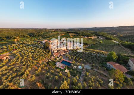 Ein Luftbild-Panorama bei Sonnenuntergang zeigt ein historisches toskanisches Agriturismo und Dorf. Umgeben von weitläufigen Olivenhainen und Weinbergen zeigt die Szene ein Stockfoto