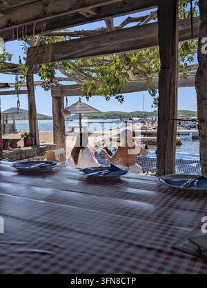 Pirovac, Kroatien 2026 Zwei Frauen sitzen an einem Strandtisch unter einer hölzernen Pergola, halten Weingläser und blicken auf die Marina mit Segelbooten, Sonnenschirm Stockfoto