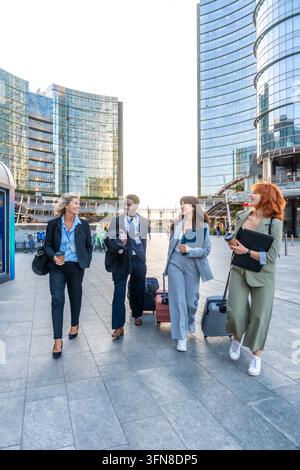 Gruppe multiethnischer Geschäftsleute, die mit Gepäck durch ein zeitgenössisches Finanzviertel gehen, lächeln und sich unterhalten, während sie geschäftlich unterwegs sind Stockfoto