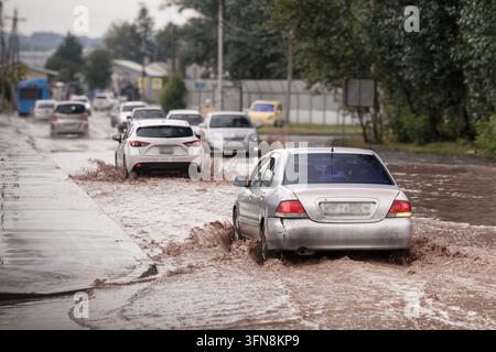 Autos fahren durch tiefes Flutwasser auf einer Stadtstraße nach starkem Regen und erzeugen große Spritzer von schlammigem Wasser. Urbane Überschwemmungen, schlechte Entwässerung, schwierig Stockfoto