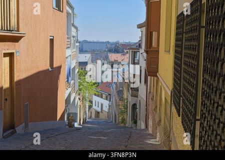 Blick den steilen Kopfsteinpflasterhang der Gül-Baba-Straße in Budapest hinunter, der farbenfrohe alte Gebäude zeigt, die eine ferne Aussicht auf Pest einrahmen. Stockfoto
