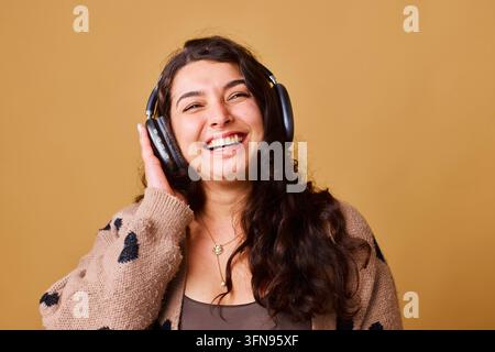 Fröhliche junge Frau hört Musik mit Kopfhörern in einem gemütlichen braunen Studio-Porträt Stockfoto
