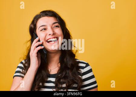 Glückliche junge Frau lacht, während sie in einem lässigen gestreiften Hemd am Smartphone spricht, vor hellem gelbem Hintergrund Stockfoto