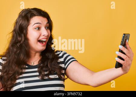 Aufgeregte Frau nimmt Selfie mit Smartphone im Studio auf gelbem Hintergrund in gestreiftem Hemd Stockfoto