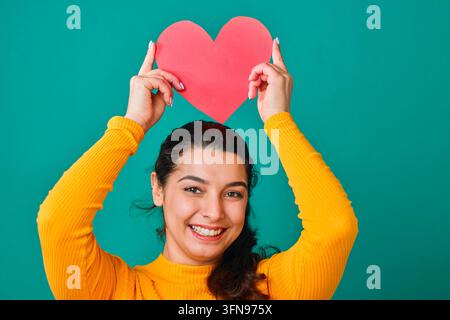 Lächelnde junge Frau hält rotes Papierherz über dem Kopf und zeigt Liebe und Glück Stockfoto