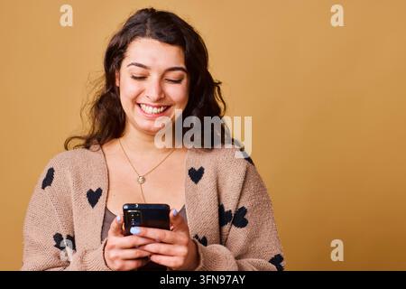 Junge Frau lächelt, während sie auf dem Smartphone schreibt, trägt eine Herz-Strickjacke und eine Goldkette Stockfoto