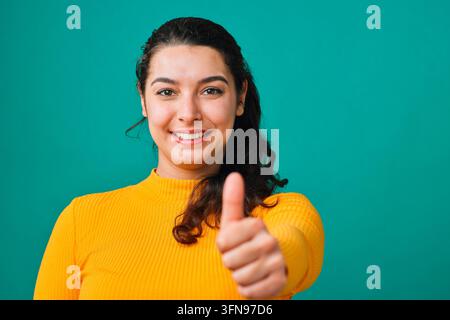 Junge Frau gibt Daumen hoch und lächelt in gelbem Pullover vor teal-farbigem Studiohintergrund Stockfoto