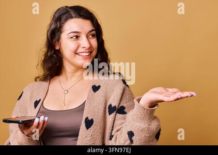 Lächelnde junge Frau, die ihre Handfläche präsentiert, während sie ein Smartphone in einem lässigen Herzen Cardigan hält, Studio-Porträt Stockfoto