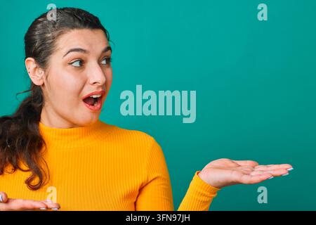 Überraschte Frau in gelbem Pullover präsentiert leere Handfläche für Produktpräsentation auf türkisfarbenem Hintergrund Stockfoto