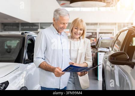 Seniorenpaar liest gemeinsam eine Fahrzeugbroschüre, vergleicht Autos in einem modernen Autohaus Stockfoto